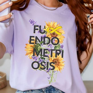 Peut inclure: T-shirt lilas avec l'inscription "F*ck Endometriosis" en noir, avec un ruban jaune et un motif floral. Le motif comprend des tournesols et de la lavande. Le t-shirt est en matière souple.