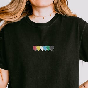 Peut inclure: T-shirt noir avec une rangée de six cœurs brodés de différentes couleurs : rose, jaune, blanc, turquoise, bleu et gris. Le t-shirt a un col rond et des manches courtes. Le tissu semble être un mélange de coton doux.