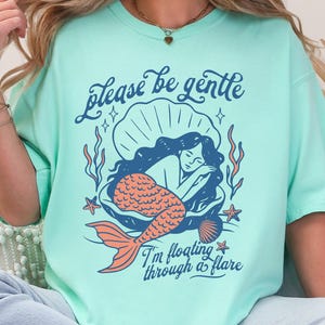 May include: A light teal t-shirt with a mermaid design inside a seashell. The mermaid has long black hair and an orange tail. The text "please be gentle" and "I'm floating through a flare" are printed on the shirt.