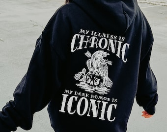 May include: Black hooded sweatshirt with white text that reads "My illness is chronic, my dark humor is iconic". The text is accompanied by a graphic of a skull with a raven perched on top.