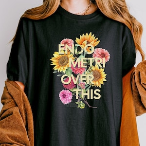 May include: Black t-shirt with the text "ENDOMETRI OVER THIS" in yellow, with a floral bouquet design. The bouquet includes sunflowers and pink flowers. The shirt is a casual style.