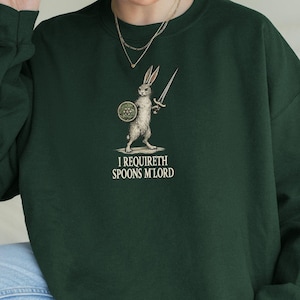 May include: Dark green sweatshirt with a rabbit warrior graphic holding a sword and shield. The text "I REQUIRETH SPOONS M'LORD" is printed below the graphic. The sweatshirt is made of a soft material.