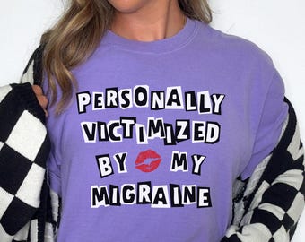 Puede incluir: Una camiseta lila con el texto "PERSONALLY VICTIMIZED BY MY MIGRAINE" en blanco, con un gráfico de labios rojos. La persona lleva vaqueros azul claro y una rebeca a cuadros en blanco y negro.