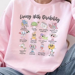 May include: A light pink crewneck sweatshirt featuring the text Living With Disability and a grid of twelve colorful cartoon illustrations with captions including checking accessibility, pacing myself, plans pending symptoms, and mobility aid freedom.