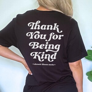 Puede incluir: Una camiseta negra de manga corta con texto blanco en la espalda que dice Thank You for Being Kind con el texto más pequeño +chronic illness sucks+ debajo. La camiseta se combina con pantalones cortos de mezclilla azul claro con dobladillos deshilachados.