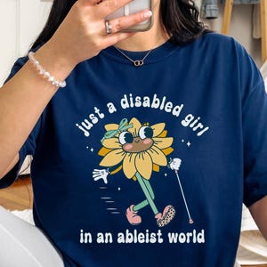 Könnte beinhalten: Marineblaues T-Shirt mit dem Text "just a disabled girl in an ableist world" und einer Cartoon-Sonnenblumenfigur mit einem Gehstock. Die Sonnenblume ist gelb mit einem grünen Stiel und Blättern. Das T-Shirt hat einen lässigen Stil.