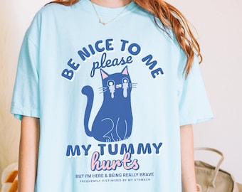 Puede incluir: Camiseta azul claro con un gato de dibujos animados llorando y el texto "Sé amable conmigo por favor, me duele la barriga. Pero estoy aquí y siendo realmente valiente, frecuentemente víctima de mi estómago."