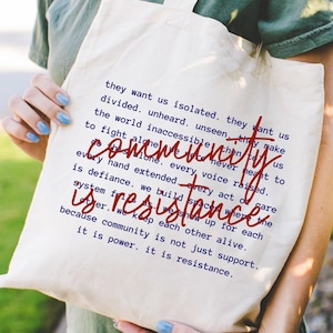 May include: A cream-colored canvas tote bag featuring printed text in dark blue that reads, they want us isolated. they want us divided. unheard. unseen. they make the world inaccessible. they never meant us to fight alone. every voice raised is defiance. we build spaces where every hand extended is resistance. we show up for each other. we keep each other alive. because community is not just support. it is power. it is resistance.