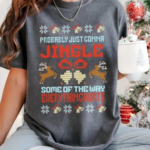 Op de afbeelding: Donkergrijs t-shirt met een feestelijk kerstontwerp. Het shirt heeft de tekst "Probably Just Comma Jingle All The Way Everything Hurts" in rood en blauw, samen met rendieren en bloemmotieven. Een gezellige vakantiekleding.