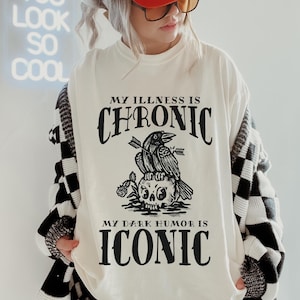 May include: A white long-sleeved shirt with black text that reads "My illness is chronic, my dark humor is iconic". The shirt features a black and white illustration of a skull with two crows perched on top. The model is wearing a red trucker hat with white text that reads "Mad love for you".