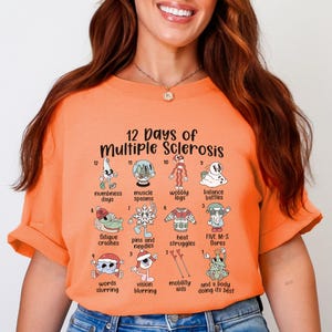May include: An orange short-sleeved t-shirt featuring a graphic print titled 12 Days of Multiple Sclerosis. The design includes twelve numbered illustrations with text labels describing symptoms like numbness, muscle spasms, and fatigue, arranged in a grid.
