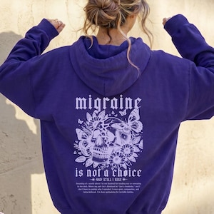 Puede incluir: Una sudadera con capucha morada con un gráfico de calavera y flores en la espalda. El gráfico incluye el texto "migraine is not a choice" y "and still I rise". La sudadera tiene una capucha con cordón y mangas largas.