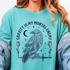 May include: Teal-colored sweatshirt with a graphic of a raven perched on a sword, surrounded by the text "GRAVITY IS MY MORTAL ENEMY". The design includes a crescent moon, stars, and floral accents. The shirt has long sleeves.