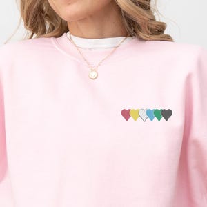 Peut inclure: Un sweat-shirt rose clair avec une rangée de six cœurs colorés brodés sur la poitrine. Les cœurs sont rouges, jaunes, bleu clair, verts et gris foncé. Le sweat-shirt a un col rond et des manches longues.