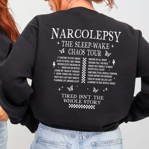 May include: A black long-sleeve sweatshirt featuring white text and graphics. The front displays THE SLEEP-WAKE CHAOS TOUR. The back features NARCOLEPSY THE SLEEP-WAKE CHAOS TOUR, a list of symptoms, butterfly icons, and TIRED ISN'T THE WHOLE STORY.
