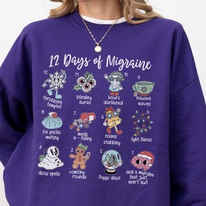 May include: A purple sweatshirt with the text "12 Days of Migraine" and illustrations depicting migraine symptoms. The illustrations include a snowflake, auras, nausea waves, and light flares. The sweatshirt is a comfortable, casual style.
