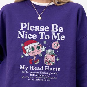 Puede incluir: Una sudadera morada con el texto "Please Be Nice To Me" y "My Head Hurts". El diseño incluye un cerebro con un gorro de Papá Noel y un tarro de caramelos de colores. La frase "FEELING A BRAIN FULL OF PAIN" también está impresa.