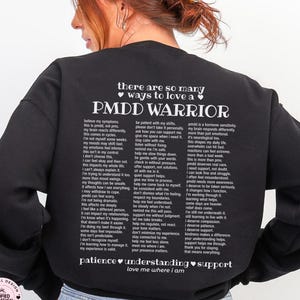 May include: Black sweatshirt with white text: "there are so many ways to love a PMDD WARRIOR." The text includes a list of supportive statements. The sweatshirt also has the words "patience, understanding, support, love me where I am." The bottom of the sweatshirt says "BLACK."