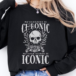 May include: Black long-sleeve shirt with the text "MY HEADACHE IS CHRONIC, MY DARK HUMOR IS ICONIC" in white. The graphic includes a skull, roses, and decorative flourishes. The shirt is a casual wear item.