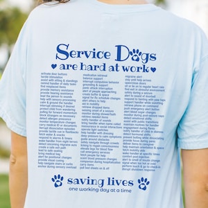 Puede incluir: Camiseta azul claro con el texto azul marino "Service Dogs are hard at work". La camiseta tiene una lista de tareas que realizan los perros de servicio. La parte inferior de la camiseta dice "saving lives one working day at a time" en azul marino.