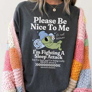Könnte beinhalten: Ein anthrazitfarbenes T-Shirt mit einer Grafik eines grünen Wesens mit blauer Mütze, das auf einer blauen Decke liegt. Der Text lautet: Please Be Nice To Me, it's not laziness, I'm Fighting A Sleep Attack, but I'm here and I'm being really BRAVE about it, NARCOLEPSY AWARENESS. Eine mehrfarbige, grob gestrickte Strickjacke wird über dem T-Shirt getragen.