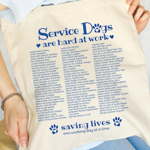 Puede incluir: Bolso tote de lona color crema con texto azul que dice "Service Dogs are hard at work" y una lista de tareas. La frase "saving lives one working day at a time" también está impresa en la bolsa.