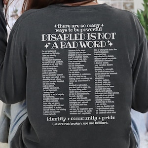 May include: A charcoal gray long-sleeve sweatshirt featuring white text on the back that reads: there are so many ways to be powerful, DISABLED IS NOT A BAD WORD, followed by a list of empowering statements and the phrase identity + community + pride.