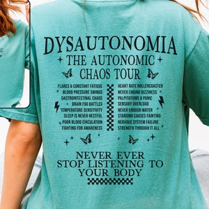 May include: A teal t-shirt with black text. The front reads "THE AUTONOMIC CHAOS TOUR". The back features the words "DYSAUTONOMIA + THE AUTONOMIC + CHAOS TOUR" with a list of symptoms and the phrase "NEVER EVER STOP LISTENING TO YOUR BODY".