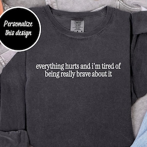 May include: Charcoal grey long-sleeved sweatshirt with the white text "everything hurts and I'm tired of being really brave about it". The sweatshirt is displayed with denim and a denim jacket. A circle with the text "Personalize this design" is in the upper left corner.