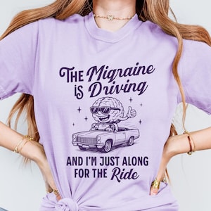 May include: A lilac t-shirt featuring the text "The Migraine is Driving" and "And I'm Just Along For The Ride". The graphic depicts a brain in sunglasses driving a car. The shirt is made of a soft, comfortable material.