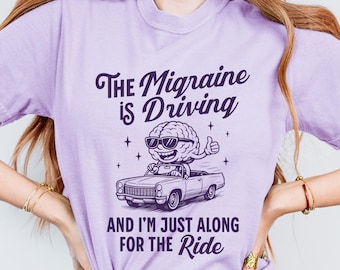 Puede incluir: Una camiseta de color lila claro con el texto "The Migraine is Driving" y "And I'm Just Along For The Ride". El gráfico muestra un cerebro con gafas de sol conduciendo un descapotable. La camiseta es de un material suave y cómodo.