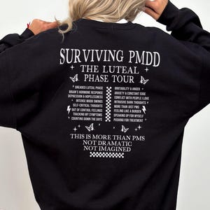May include: Black sweatshirt with white text: "SURVIVING PMDD THE LUTEAL PHASE TOUR." The back of the sweatshirt lists symptoms and phrases related to PMDD. The bottom of the sweatshirt reads "THIS IS MORE THAN PMS NOT DRAMATIC NOT IMAGINED."