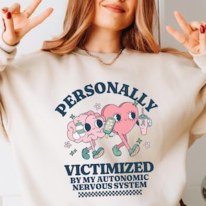 Puede incluir: Una sudadera de color crema con un diseño gráfico que presenta un cerebro y un corazón sosteniendo bebidas. El texto dice "PERSONALLY VICTIMIZED BY MY AUTONOMIC NERVOUS SYSTEM".