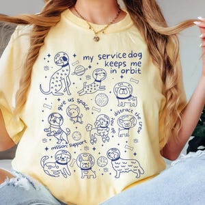 Könnte beinhalten: Hellgelbes T-Shirt mit einer Grafik von Hunden in Raumanzügen und dem Text "my service dog keeps me in orbit". Das Design umfasst Planeten, Knochen und die Sätze "give us space", "distract the crew" und "mission support".