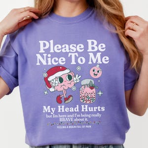 Puede incluir: Una sudadera lila con el texto "Please Be Nice To Me" y "My Head Hurts". El gráfico presenta un cerebro de dibujos animados con un gorro de Papá Noel y un tarro de bayas rosas. También se incluye el texto "Feeling a brain full of pain".