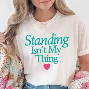 Könnte beinhalten: Ein cremefarbenes Kurzarm-T-Shirt mit dem türkisfarbenen Schriftzug "Standing Isn't My Thing" mittig auf der Brust über einem kleinen rosa Herz. Das T-Shirt wird mit einer blauen Jeans und einer grob gestrickten Strickjacke mit rosa und blauen Streifen getragen.