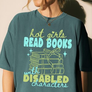Pode incluir: Uma t-shirt de manga curta em verde-azulado escuro com um gráfico de uma pilha de livros atados com uma fita. O texto hot girls READ BOOKS with DISABLED characters está escrito em verde claro e azul vivo com estrelas.