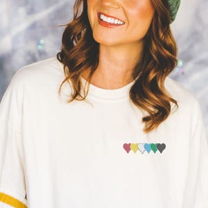 Peut inclure: T-shirt blanc cassé orné d'une rangée de six cœurs brodés de différentes couleurs. Les cœurs sont disposés en ligne, avec des couleurs telles que le rouge, le jaune, le vert et le gris. Le t-shirt a une coupe ample et des manches courtes.