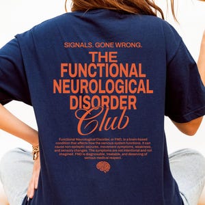 Op de afbeelding: Marineblauw T-shirt met oranje tekst: "SIGNALS, GONE WRONG. THE FUNCTIONAL NEUROLOGICAL DISORDER Club." Het shirt bevat ook een beschrijving van functionele neurologische stoornis. De persoon draagt een lichtblauwe jeans.