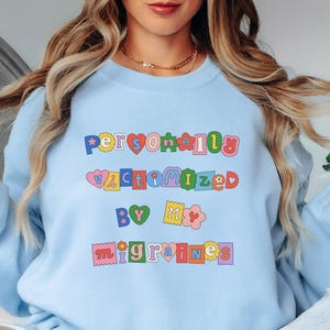 Puede incluir: Sudadera azul claro con el texto "Personalmente victimizado por mis migrañas" en letras coloridas y divertidas.