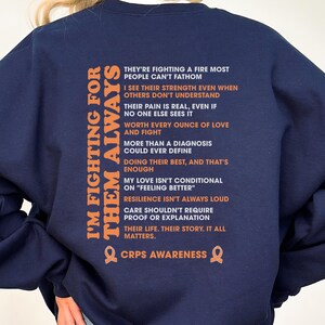 Op de afbeelding: Marineblauw sweatshirt met de oranje tekst "I'M FIGHTING FOR THEM ALWAYS". De achterkant toont een lijst met zinnen in oranje en de woorden "CRPS AWARENESS" onderaan. Een comfortabel, casual kledingstuk.