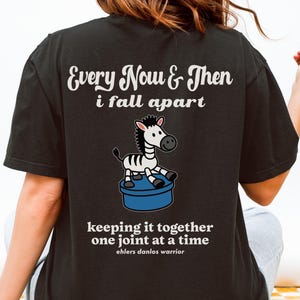 May include: Black t-shirt featuring the text "Every Now & Then i fall apart" above a cartoon zebra on a blue cylinder. The text "keeping it together one joint at a time" and "ehlers danlos warrior" is below.
