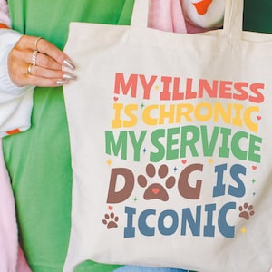 May include: A cream-colored canvas tote bag featuring multi-colored text that reads: MY ILLNESS IS CHRONIC MY SERVICE DOG IS ICONIC. The design includes brown paw prints and small star and heart accents in various colors printed on the front.