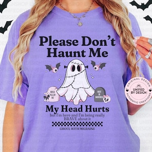 Puede incluir: Una camiseta de color lila claro con el texto "Please Don't Haunt Me" y un gráfico de fantasma. La camiseta también presenta el texto "My Head Hurts" y "Ghoul with Migraine". También se muestran murciélagos y lápidas.