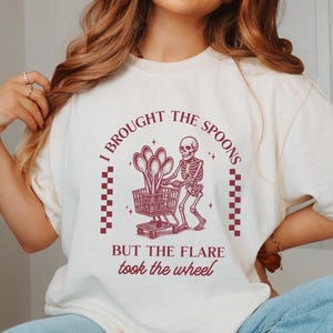 Könnte beinhalten: Cremefarbenes T-Shirt mit burgunderfarbener Grafik. Die Grafik zeigt ein Skelett, das einen Einkaufswagen mit Löffeln schiebt, mit dem Text "I BROUGHT THE SPOONS BUT THE FLARE took the wheel."