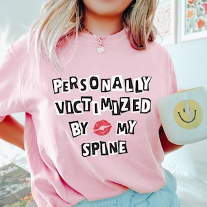 May include: Pink t-shirt with the text "PERSONALLY VICTIMIZED BY MY SPINE" in black letters with a red lip print between "BY" and "MY".