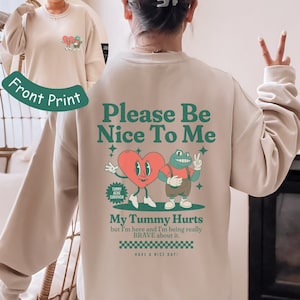 Op de afbeelding: Beige sweatshirt met een groene en rode grafische print van een hart en een kikker. De tekst "Please Be Nice To Me" is boven de afbeelding gedrukt. De tekst "My Tummy Hurts but I'm here and I'm being really BRAVE about it. HAVE A NICE DAY!" is onder de afbeelding gedrukt.