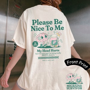 Retro migraine t-Shirt. The front has a pocket design that says Migraine Survivor with a pink brain character and a pill bottle with pills that are pastel and flower shaped. The back includes the graphic and, Please be nice to me, my head hurts