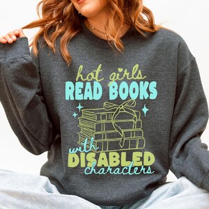 May include: A dark heather gray crewneck sweatshirt featuring a graphic print with the text "hot girls READ BOOKS with DISABLED characters" in light green and blue, accompanied by an illustration of a stack of books tied with a ribbon.