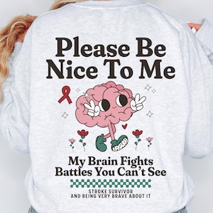 Op de afbeelding: Lichtgrijs sweatshirt met de tekst "Please Be Nice To Me" en "My Brain Fights Battles You Can't See". Het ontwerp bevat een roze hersenkarakter, een rood lint, bloemen en sterren. De woorden "STROKE SURVIVOR AND BEING VERY BRAVE ABOUT IT" zijn ook opgenomen.